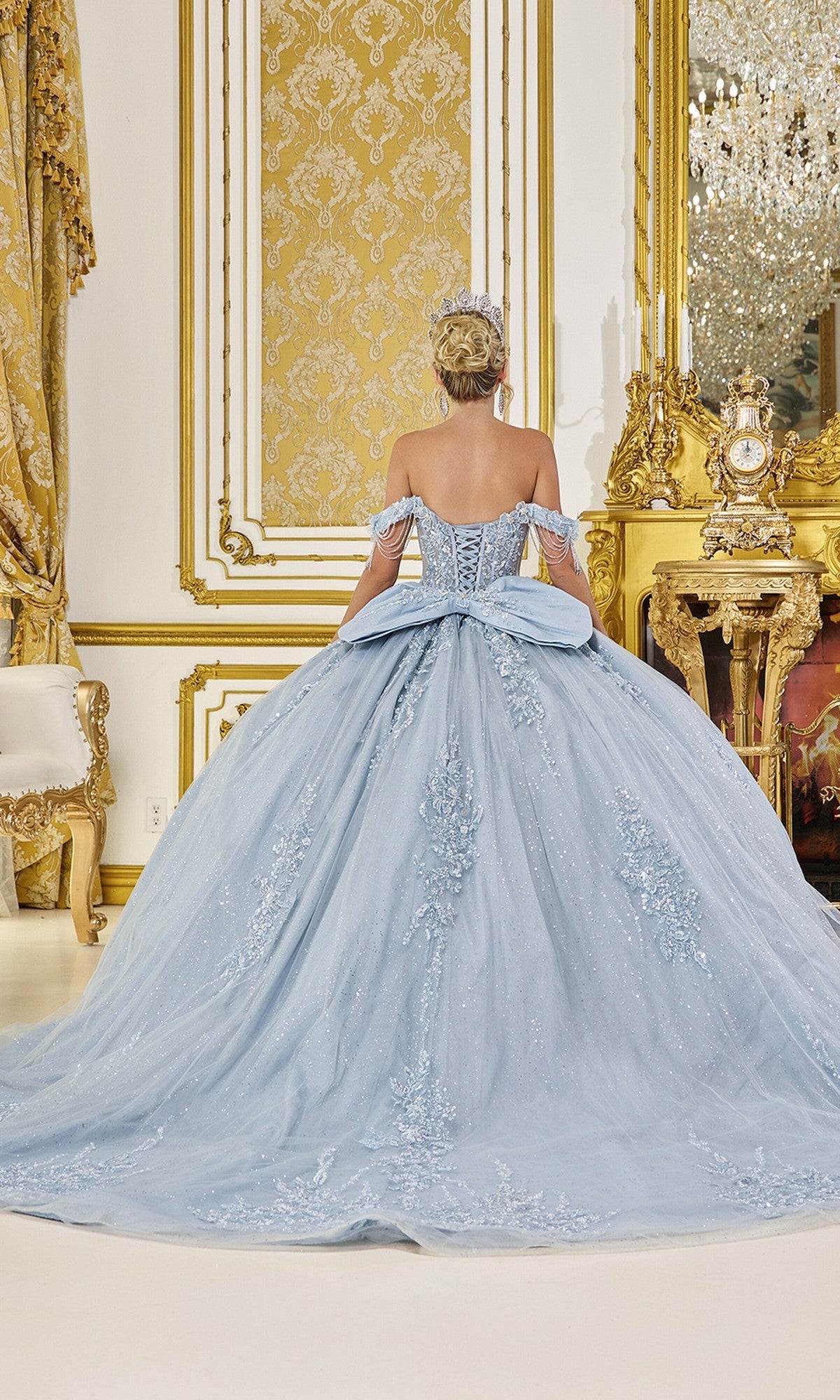A woman faces away in an ornate, gold-accented room wearing the Quinceanera Dress 5092 By Dancing Queen, a light blue ball gown with intricate embroidered applique.